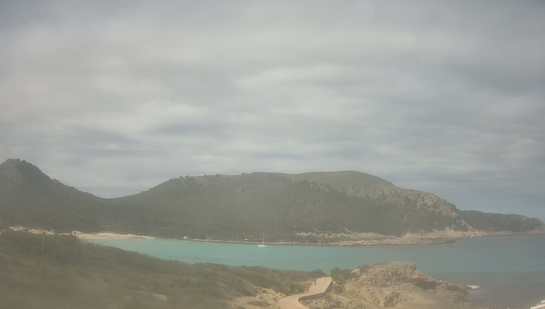 A secluded cove with turquoise water is nestled between low, green hills under a cloudy sky.