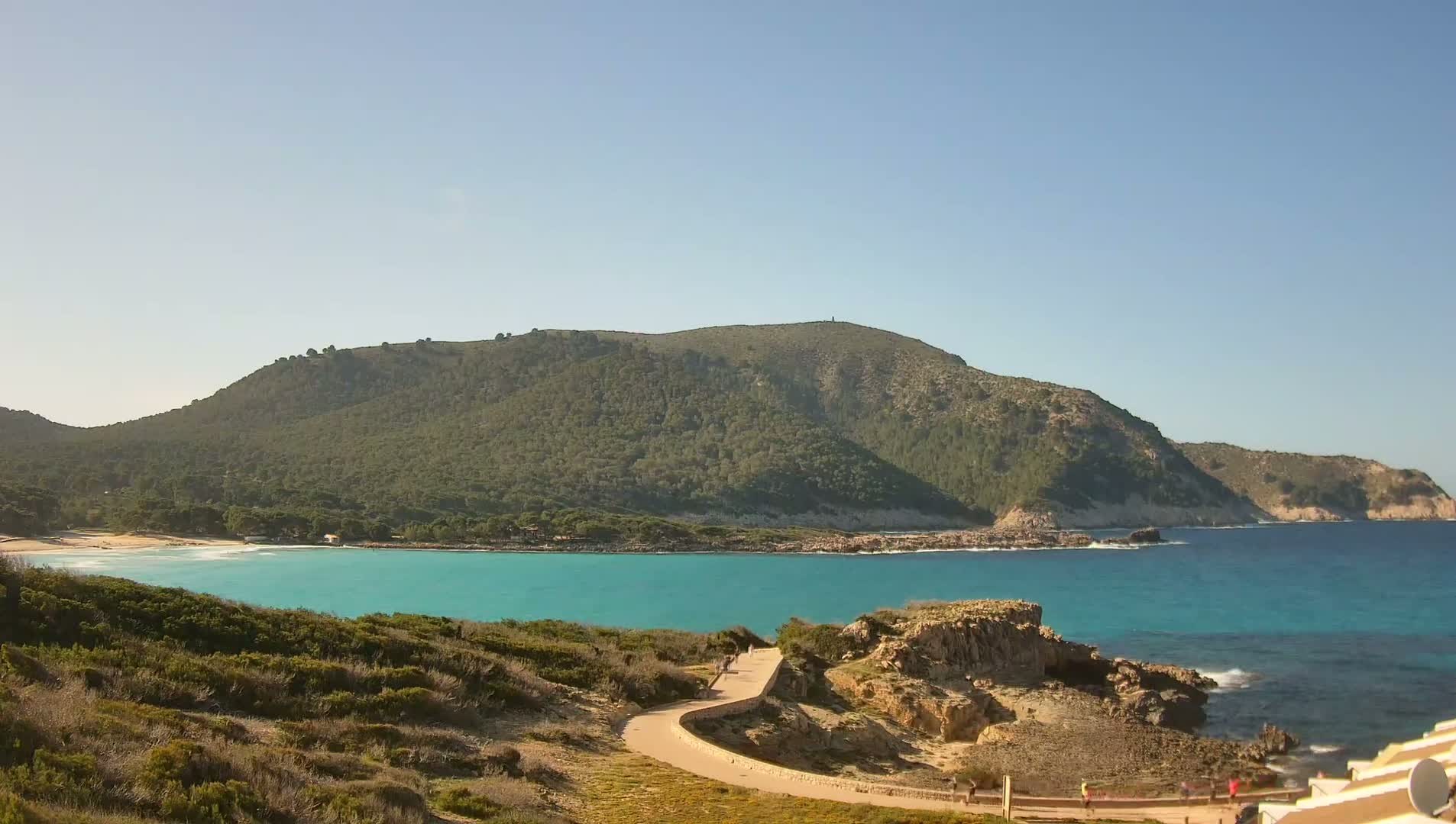 A paved path winds down a hill towards a turquoise bay with a sandy beach, backed by a verdant mountain under a clear blue sky.
