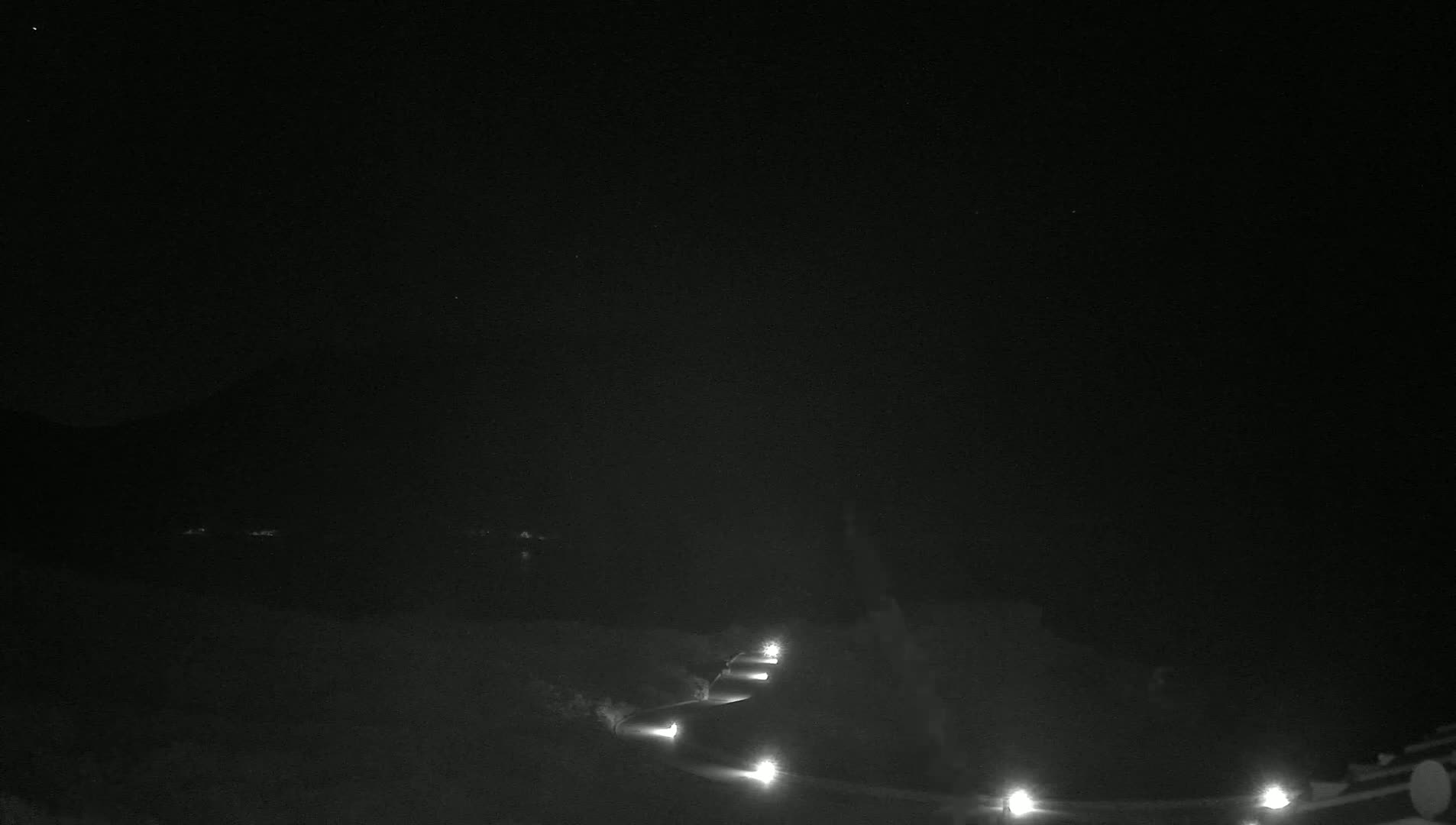 A nighttime, low-light view shows a winding road lit by several lights, leading toward a dark, mountainous landscape under a clear, starlit sky.