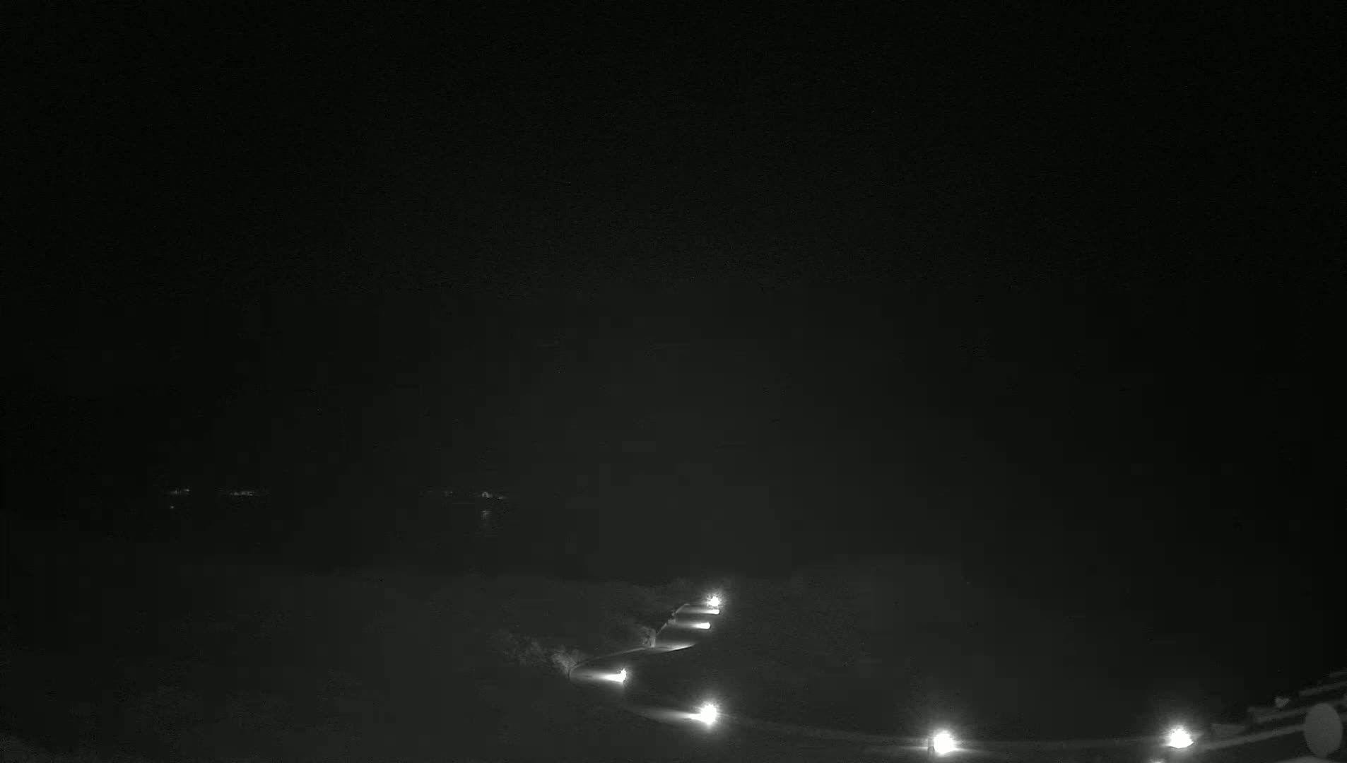 A nighttime view shows a dark, curving road illuminated by spaced lights, with a dark body of water and distant lights visible in the background.