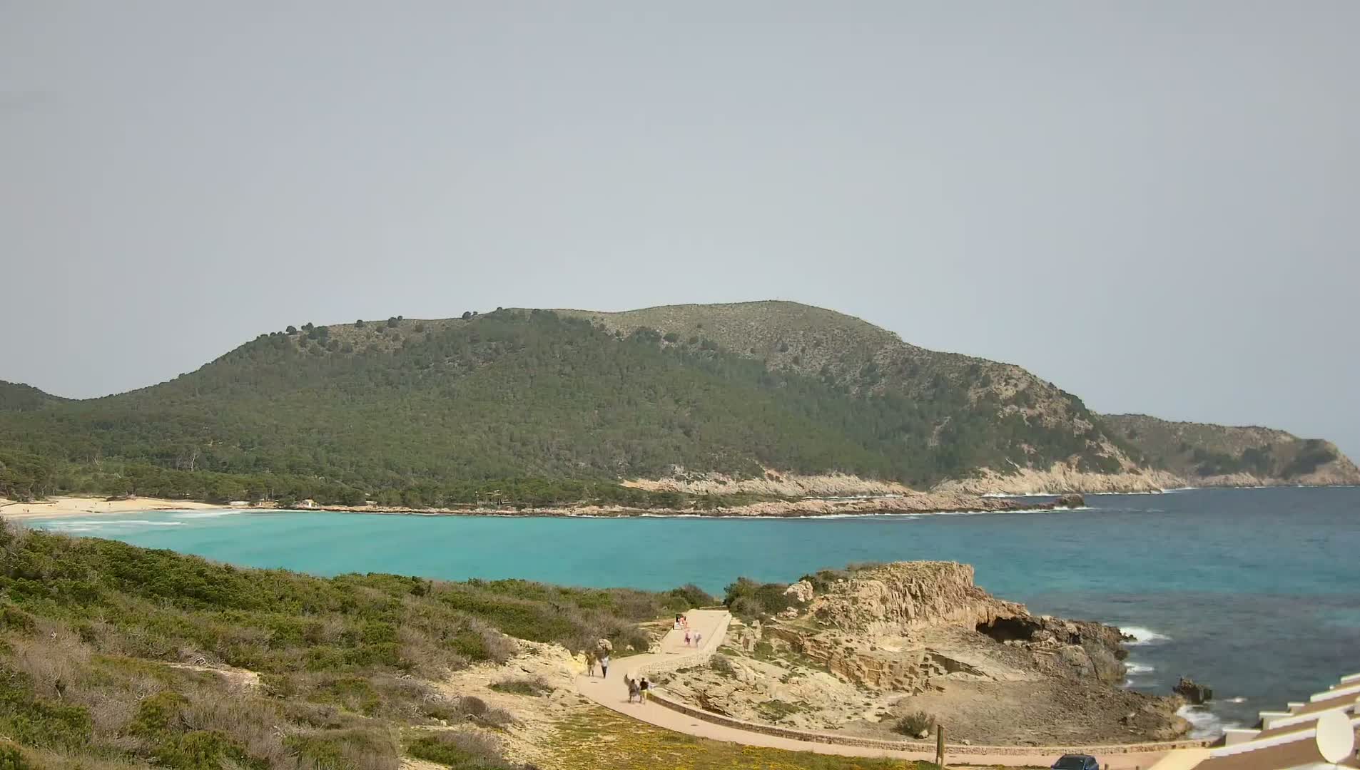 A coastal scene shows a turquoise bay, a sandy beach, a green hill, and a paved path with people walking, under a hazy sky.