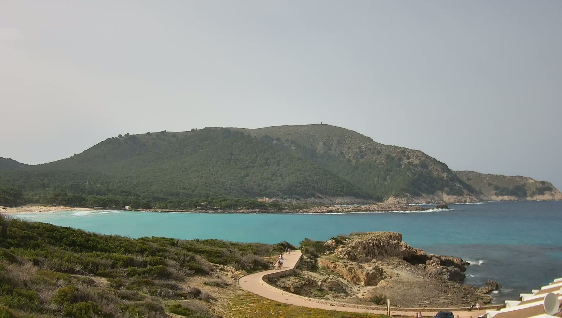 A paved path winds down a rocky hillside toward a turquoise bay, with a sandy beach and a verdant hill in the background under a bright, clear sky.