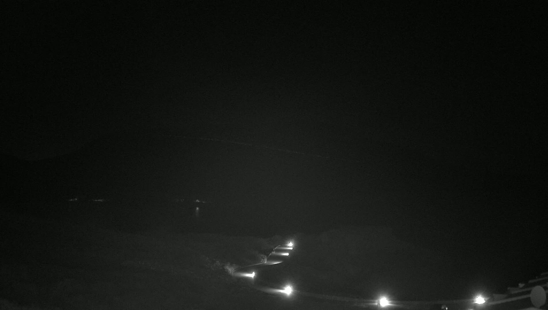 A dark, nighttime scene shows a winding road with several sets of vehicle headlights visible, leading toward a dark body of water under a clear sky.