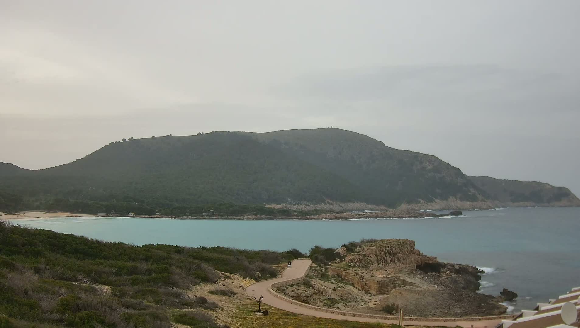 A winding road leads down to a calm, turquoise bay nestled between a verdant hill and rocky coastline under an overcast sky.