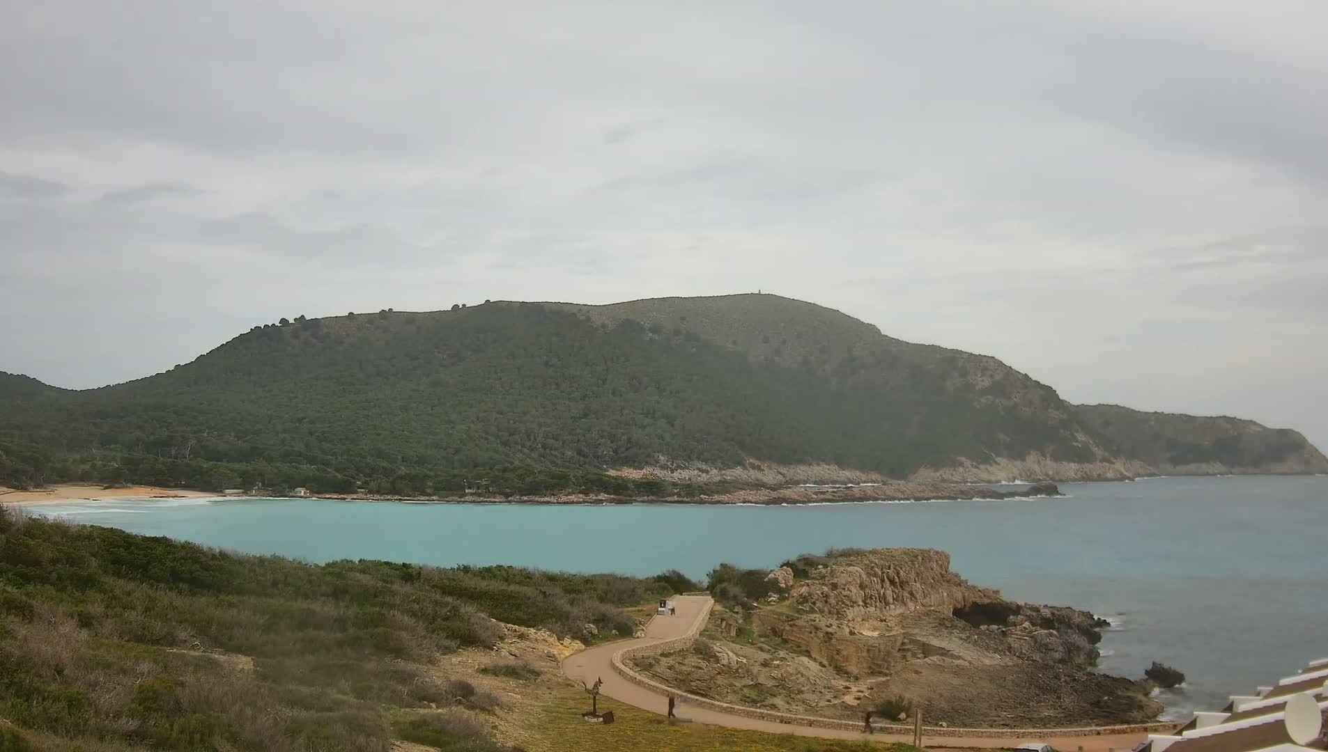 A winding road leads down to a secluded turquoise bay nestled between a verdant hill and the sea under an overcast sky.