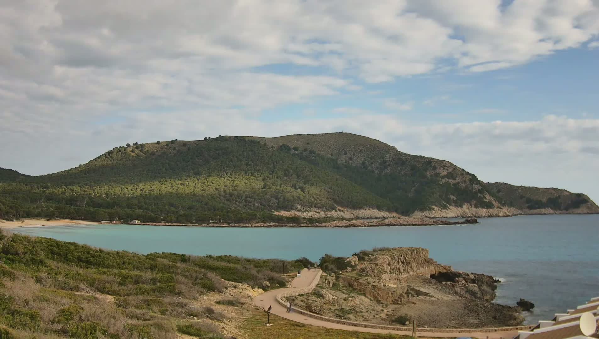 A partly cloudy sky overlooks a tranquil cove with a light teal sea, a sandy beach, and a winding path leading to a rocky headland covered in low-lying vegetation, all nestled beneath a verdant hill.