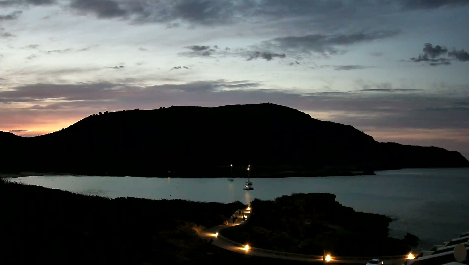 A dark, silhouetted hill overlooks a calm bay at twilight, under a mostly cloudy sky with a hint of pink and orange near the horizon.