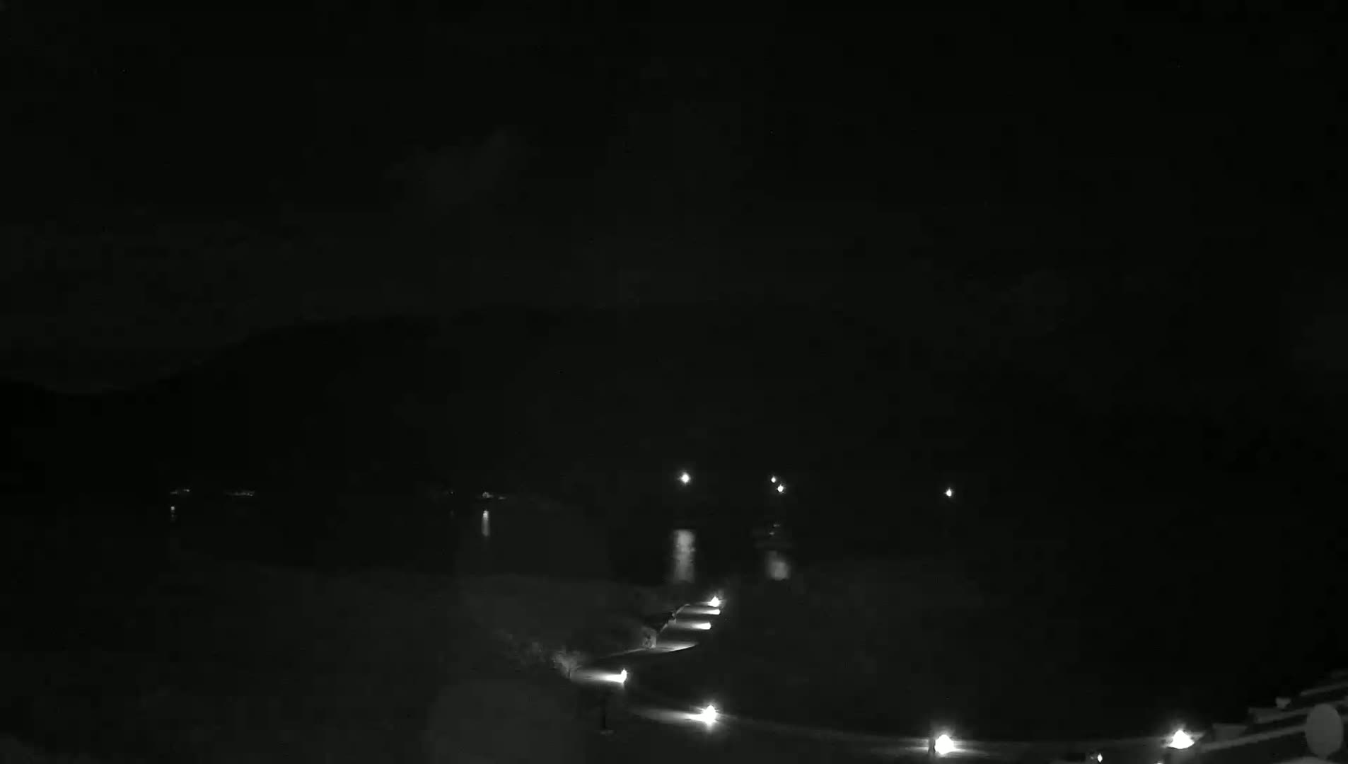 A dark nighttime scene shows a dimly lit path curving toward a body of water under a cloudy sky.