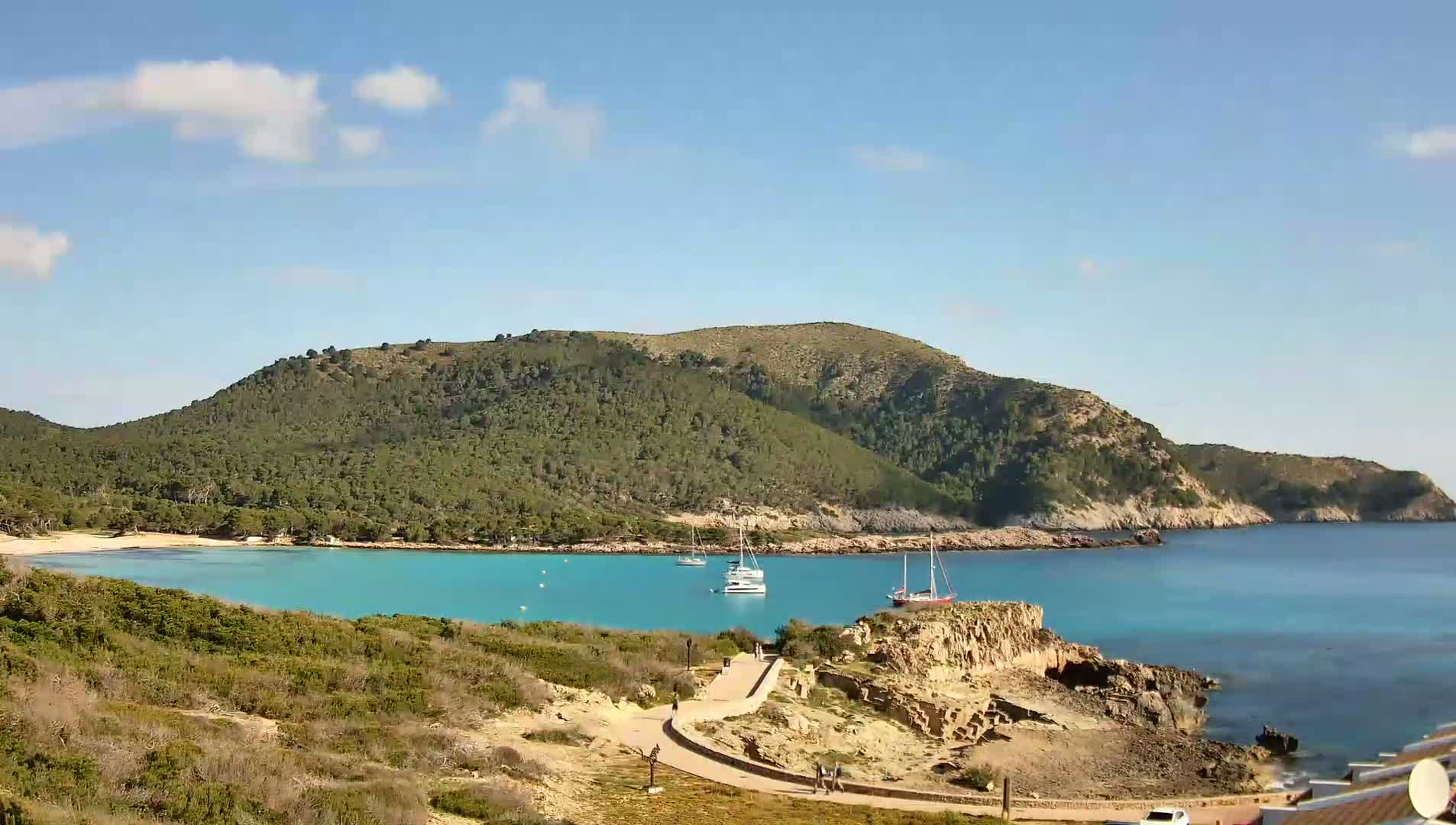 A tranquil cove with turquoise water, a sandy beach, and several boats at anchor, nestled beneath a green, hilly landscape under a sunny sky with a few scattered clouds.
