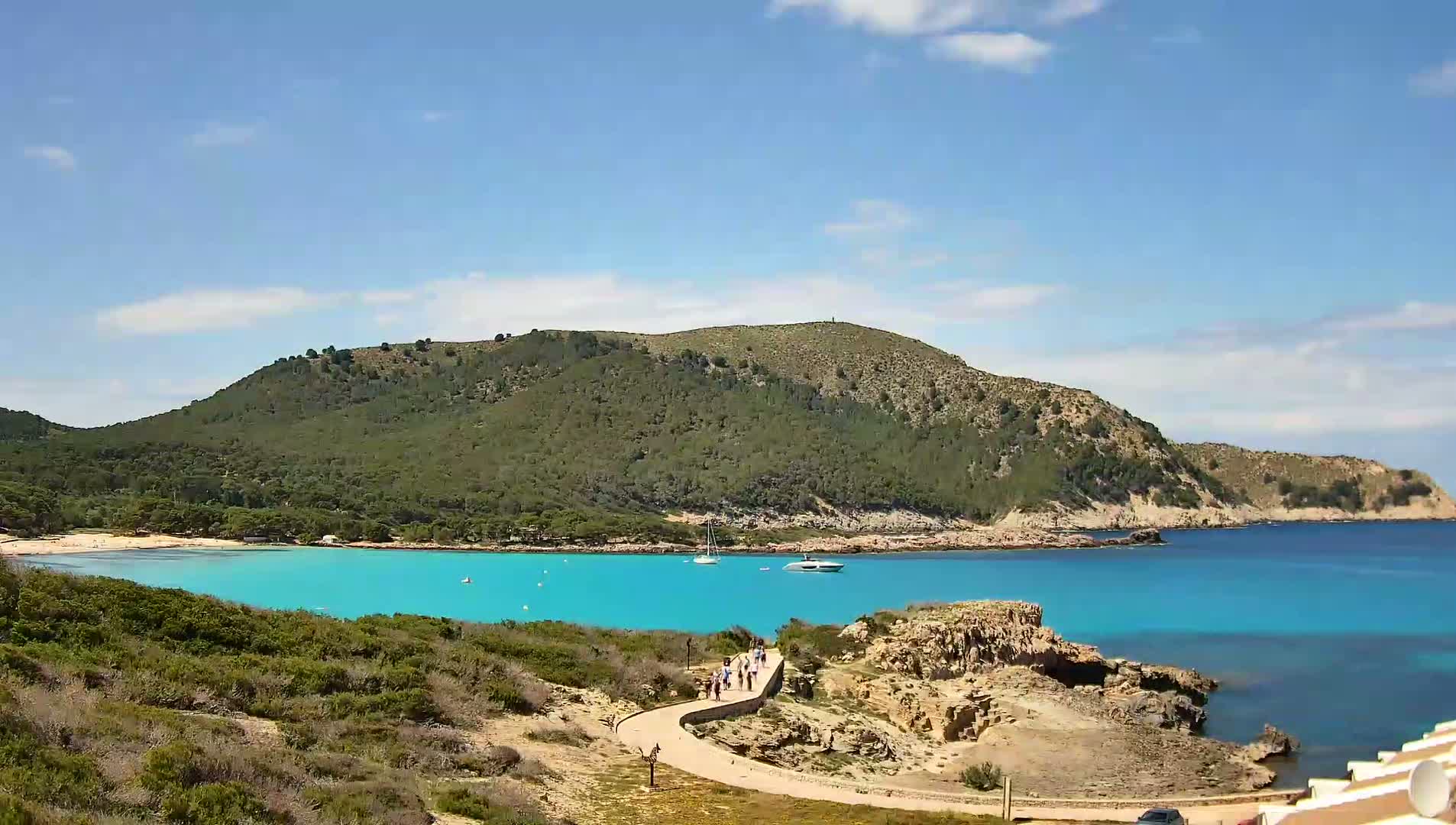 A curving path leads to a small beach with turquoise water, nestled in a cove surrounded by green hills under a mostly sunny sky.