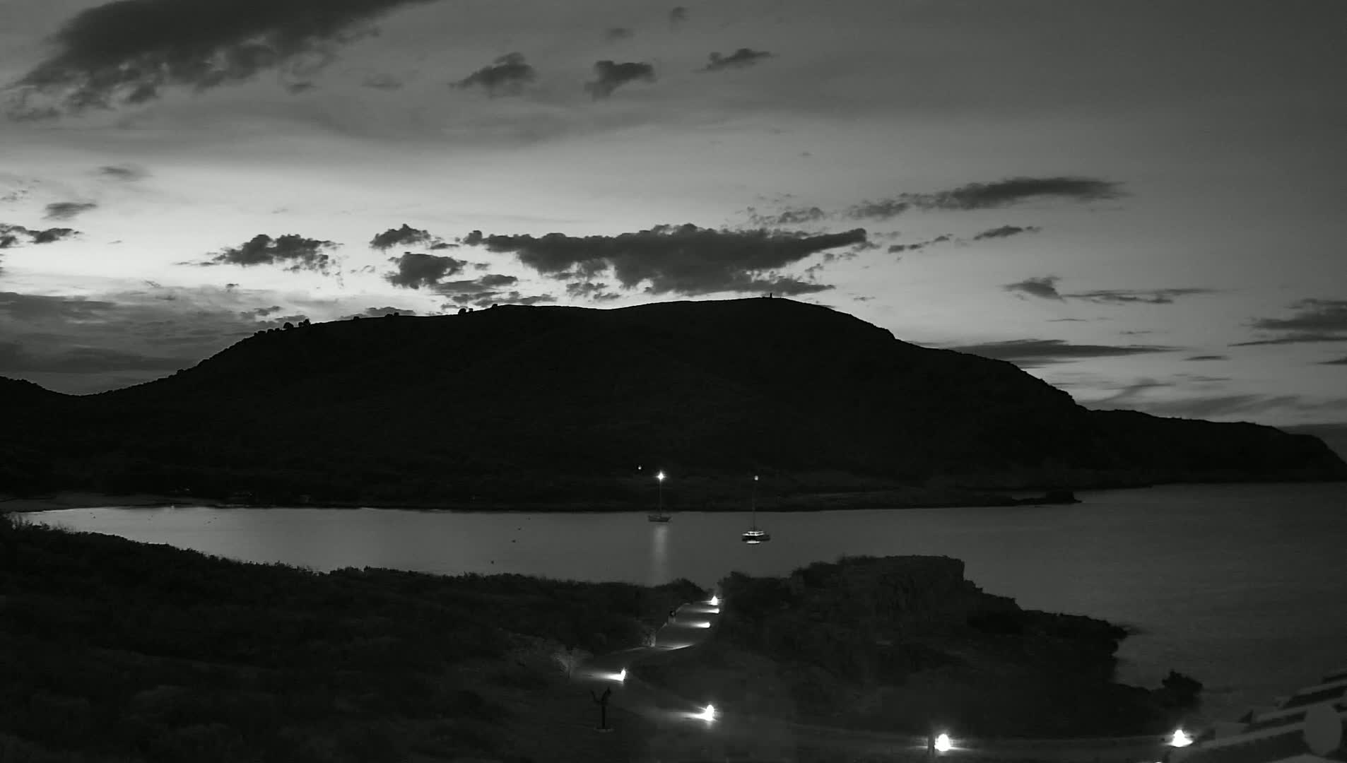 A black and white image shows a dark, silhouetted hill overlooking a calm bay at dusk under a cloudy sky, with a few lights visible along a path leading down to the water.