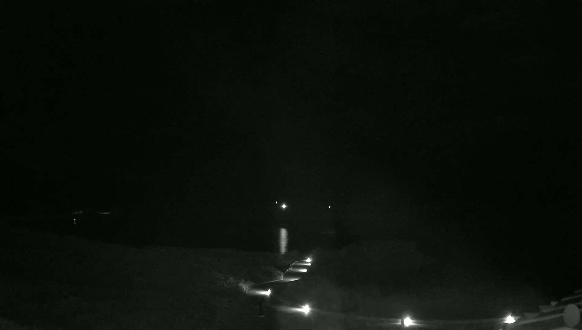 A dark, nighttime scene shows a winding road lit by several lights leading toward a body of water under a clear sky.