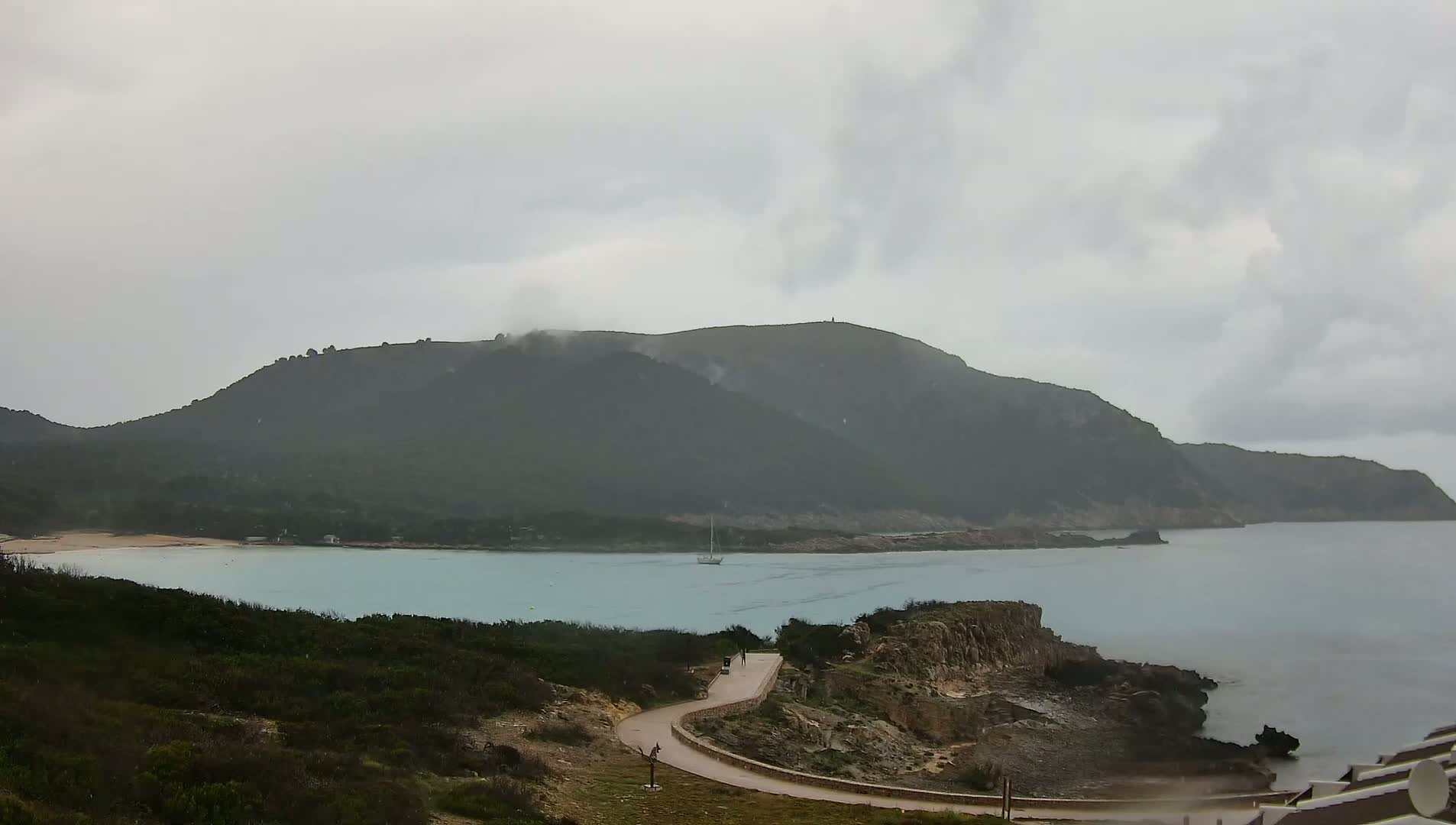 A winding road leads down to a calm, light-blue bay nestled between a low-lying, dark-green, cloud-covered mountain and a rocky coastline under an overcast sky.