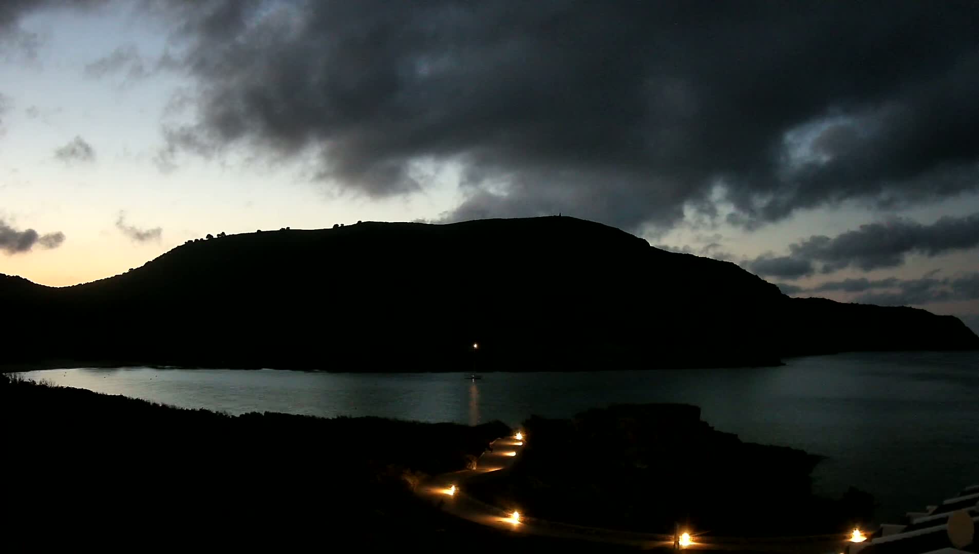 A dark, silhouetted hill overlooks a calm bay at twilight under a cloudy sky, with a path illuminated by lights winding along the shore.