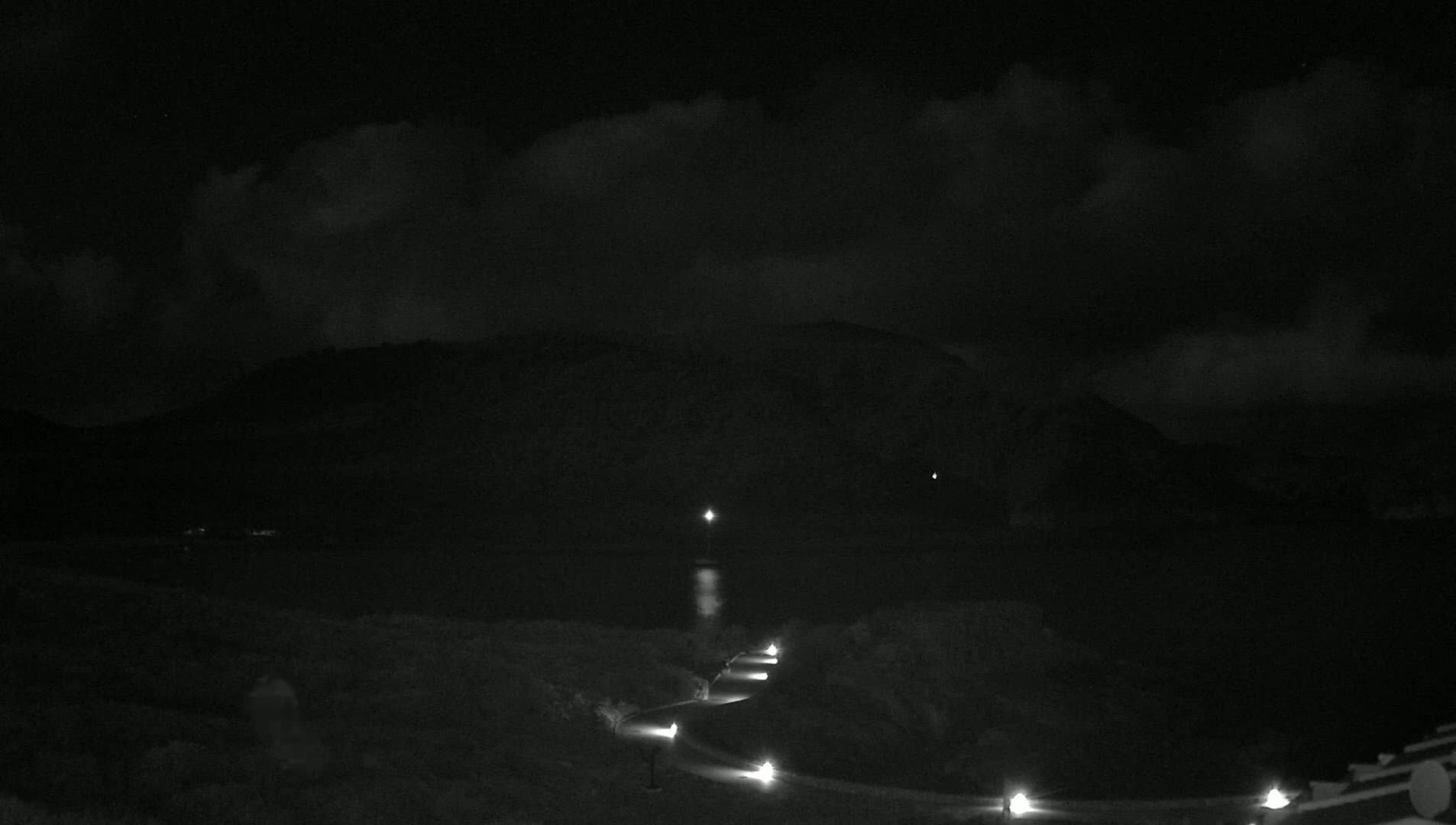 A nighttime, cloudy scene shows a winding road with lights leading down toward a dark body of water and a silhouetted mountain in the background.