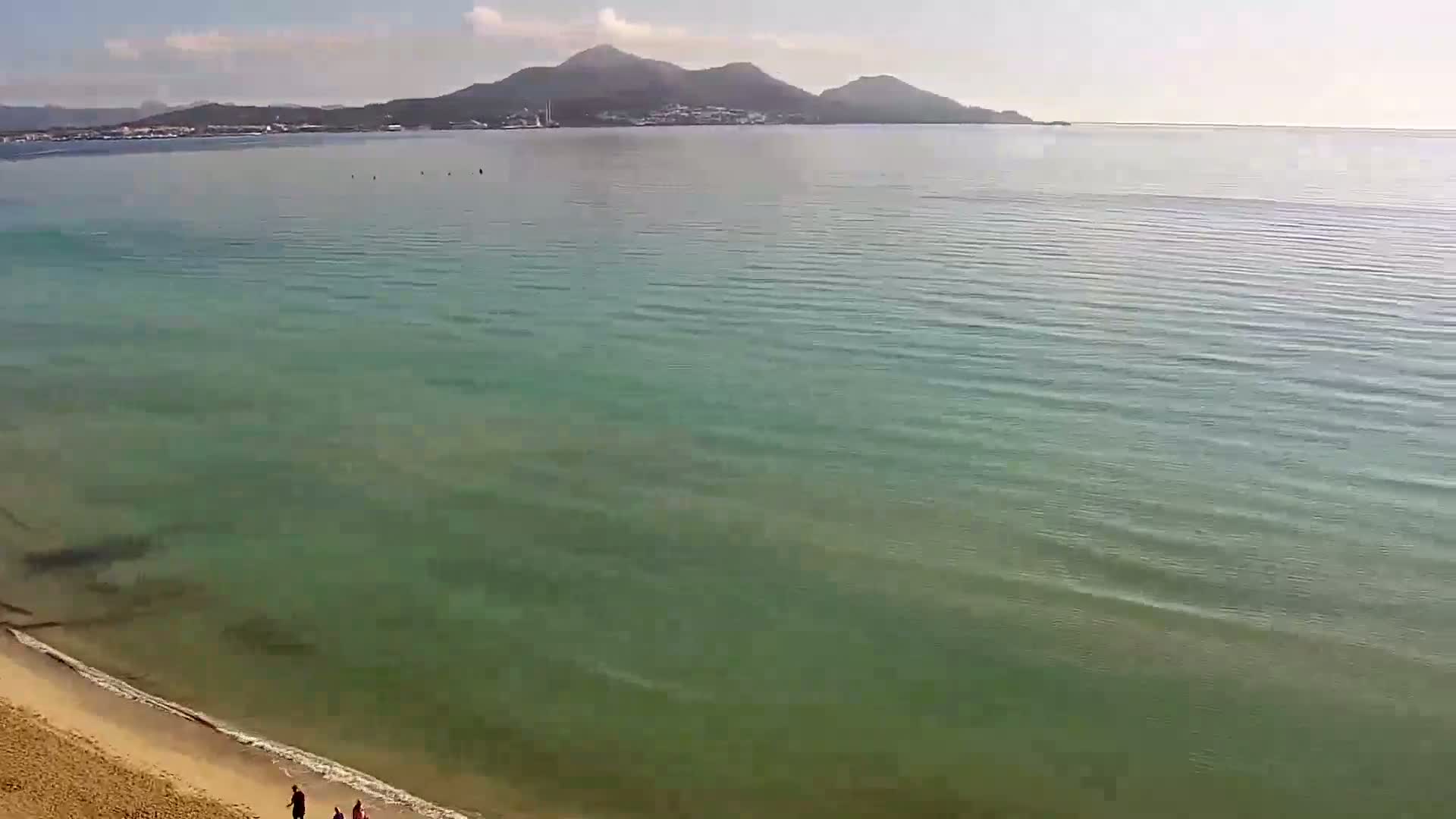 A sandy beach slopes gently into a calm, clear, teal sea, with mountains visible in the distance under a mostly sunny sky.