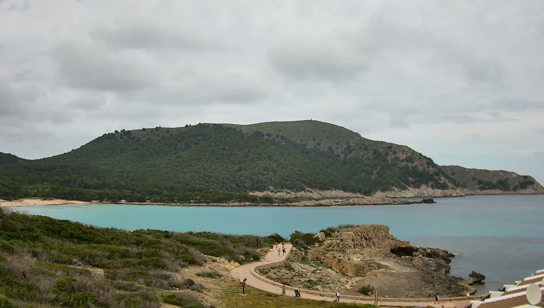 A turquoise bay is nestled between a green, hilly coastline under a cloudy sky.