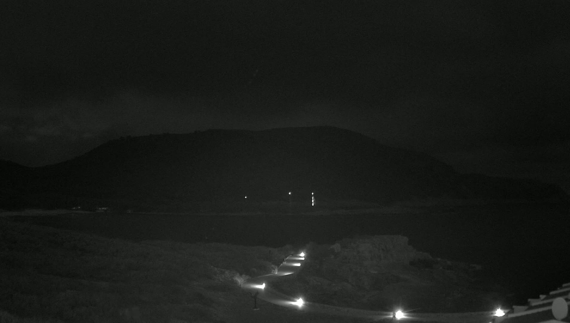 A winding road lit by lamps leads to a dark body of water under a mostly cloudy night sky, with a silhouetted hill in the background.