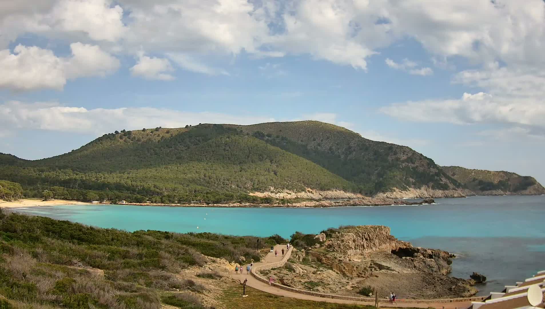 A partly cloudy sky overlooks a tranquil turquoise cove nestled between a lush green hillside and rocky shoreline, where a few people stroll along a path.
