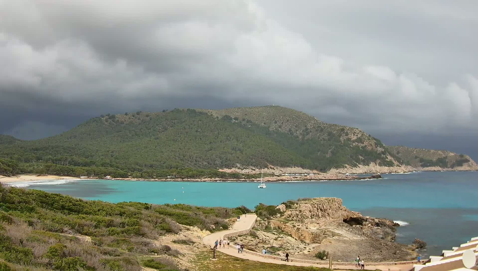 A winding path leads to a turquoise cove nestled beneath a forested hill, all under a cloudy sky.