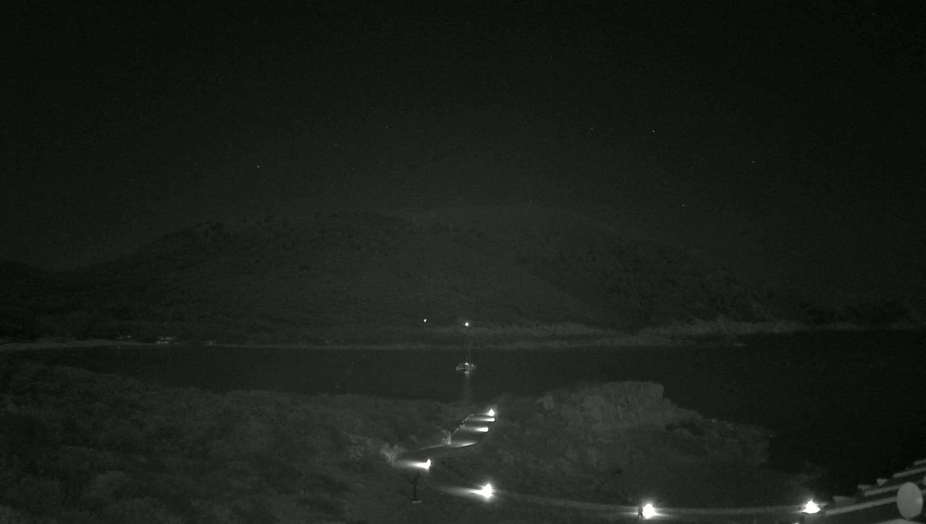 A dark, clear night scene shows a body of water with a distant hill in the background, a winding road lit by lamps in the foreground, and a small boat on the water.