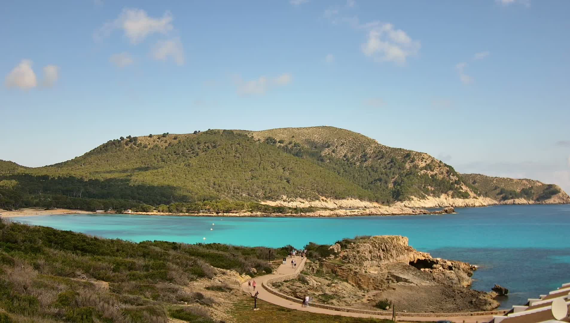 A curving paved path leads down to a turquoise bay nestled within a green, rocky hillside under a mostly clear, sunny sky.