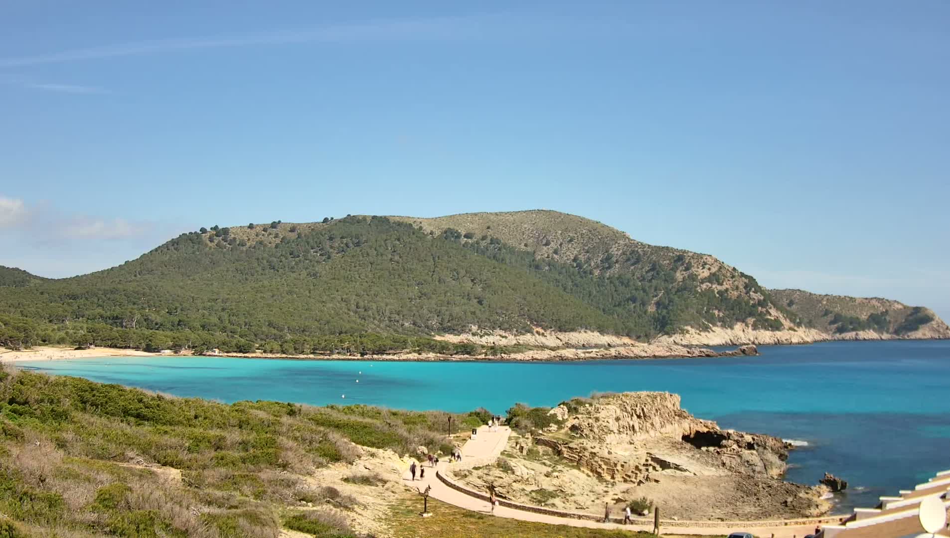 A picturesque cove with bright turquoise water, a sandy beach, and a path winding through low-lying vegetation under a clear blue sky.