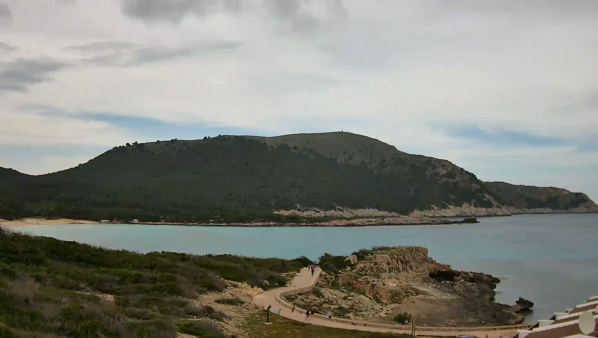 A winding path leads down to a calm, turquoise bay nestled beneath a verdant, mountainous coastline under a mostly cloudy sky.