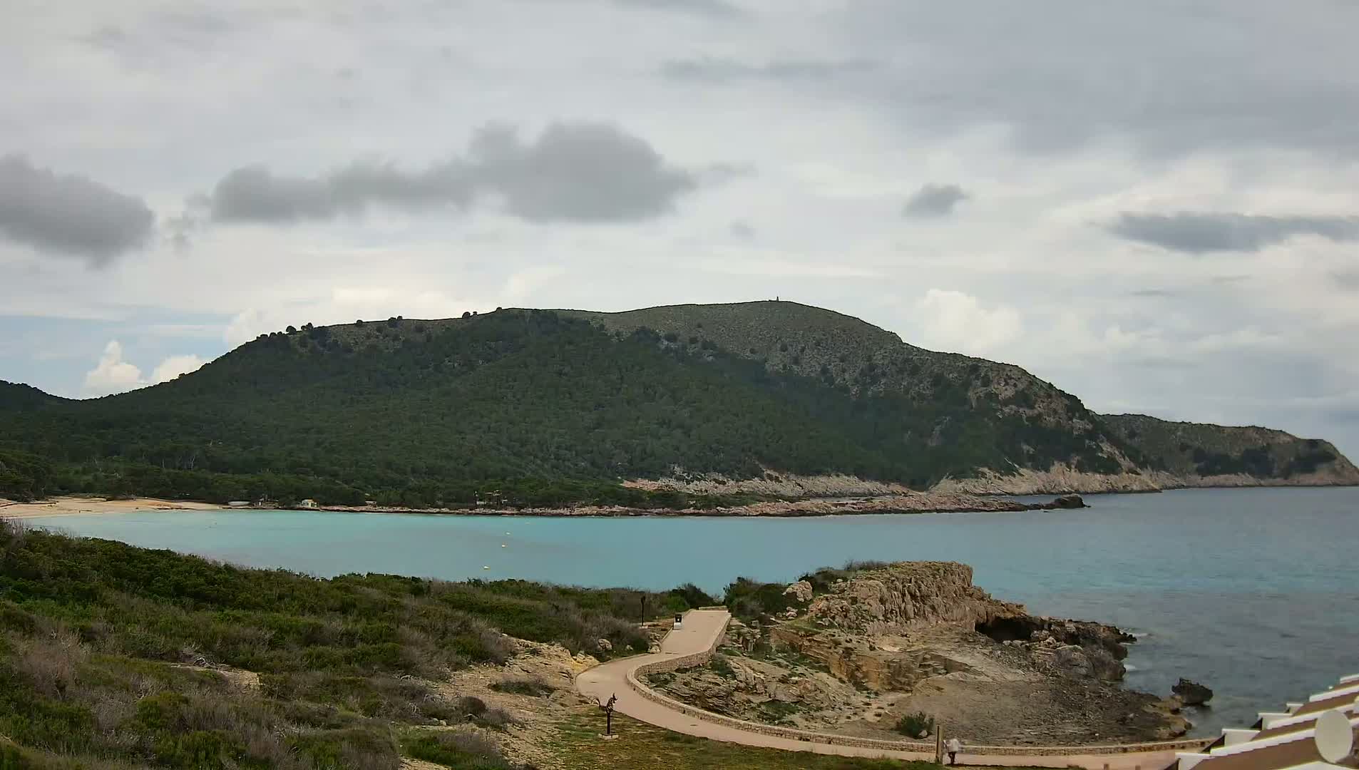 A paved path winds down toward a tranquil turquoise bay nestled below a verdant, rocky hill under a mostly cloudy sky.