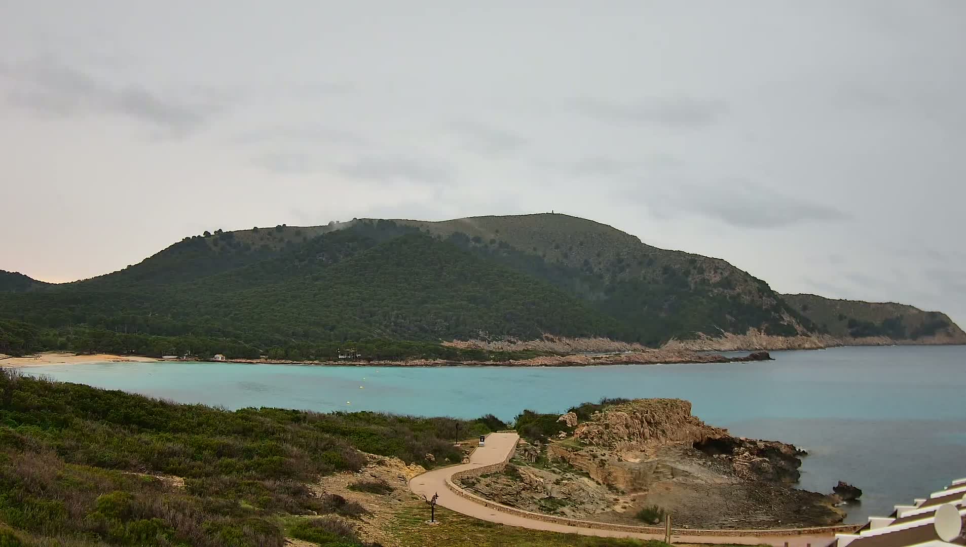 A winding path leads to a turquoise bay nestled below a verdant hill under an overcast sky.