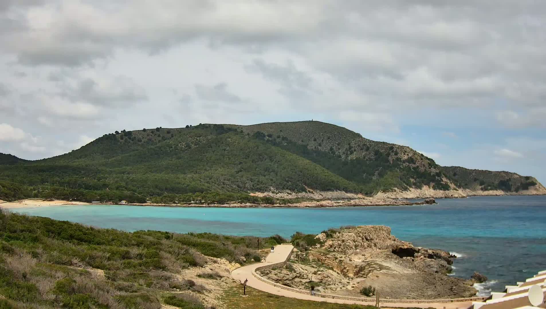 A paved path winds through coastal vegetation toward a turquoise bay, bordered by a green hill under a cloudy sky.