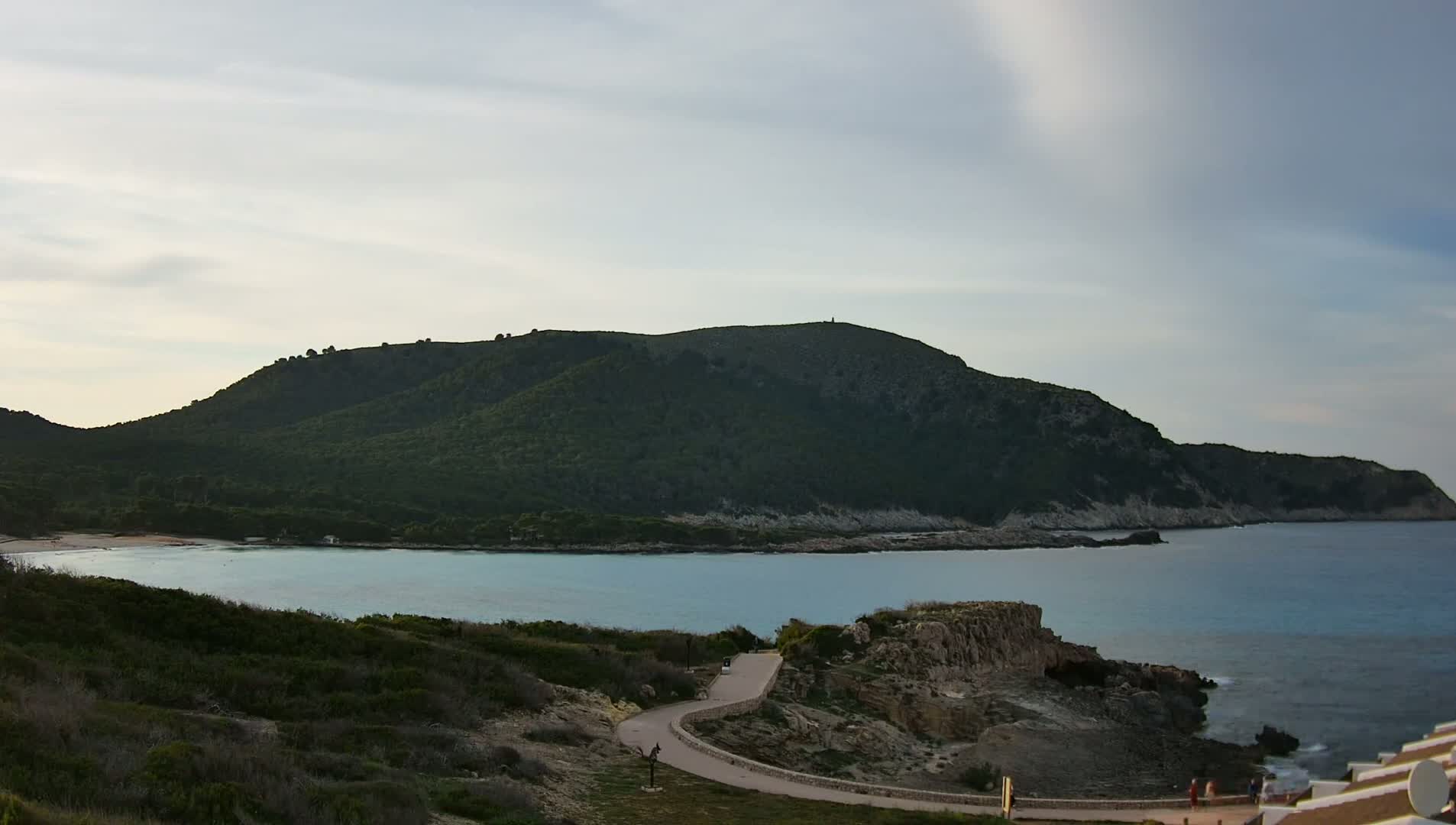A tranquil coastal scene features a calm, turquoise bay nestled between a verdant, mountainous headland and a rocky shoreline under a mostly cloudy sky.