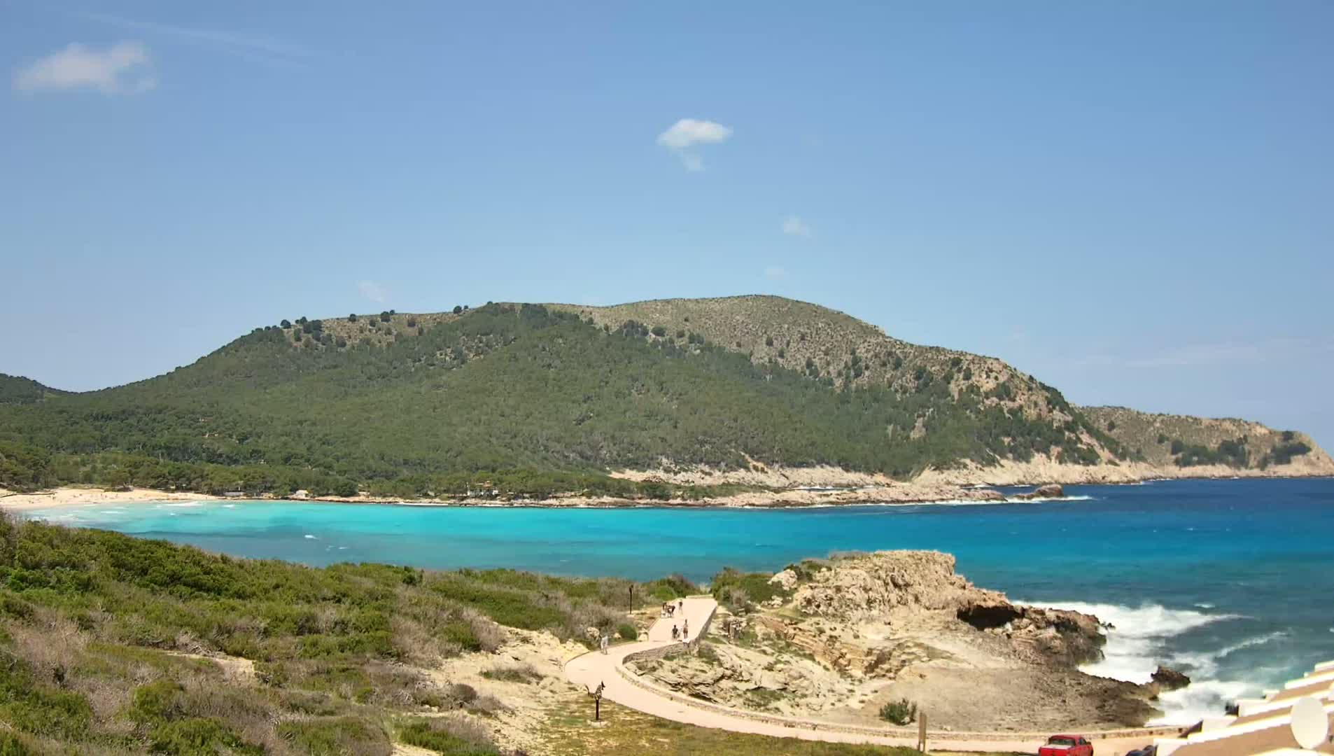 A paved path winds through coastal vegetation toward a turquoise bay bordered by a green hill under a mostly clear blue sky.
