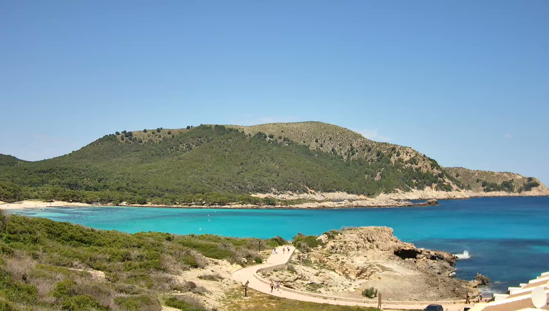 A winding path leads to a turquoise cove nestled between a verdant hill and a clear blue sea under a bright, sunny sky.