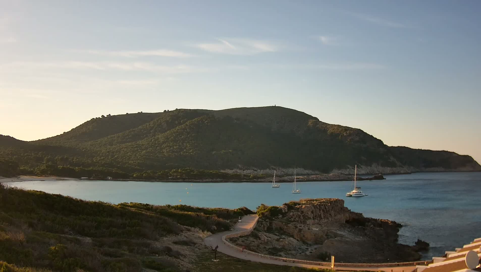 A tranquil bay with clear, turquoise water is nestled beneath a verdant hill under a mostly clear sky.
