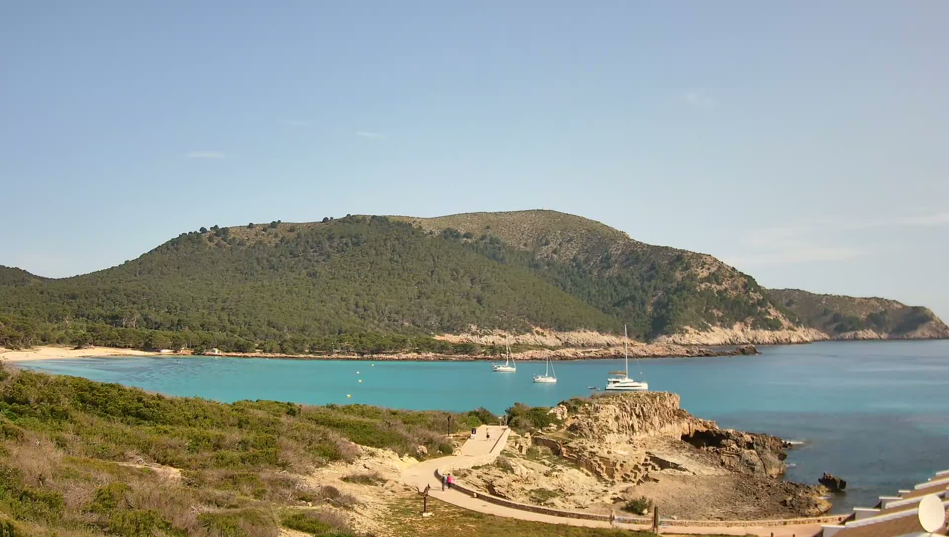 A tranquil cove with turquoise water, a sandy beach, and a few sailboats, is nestled beneath a verdant, hilly coastline on a sunny day.