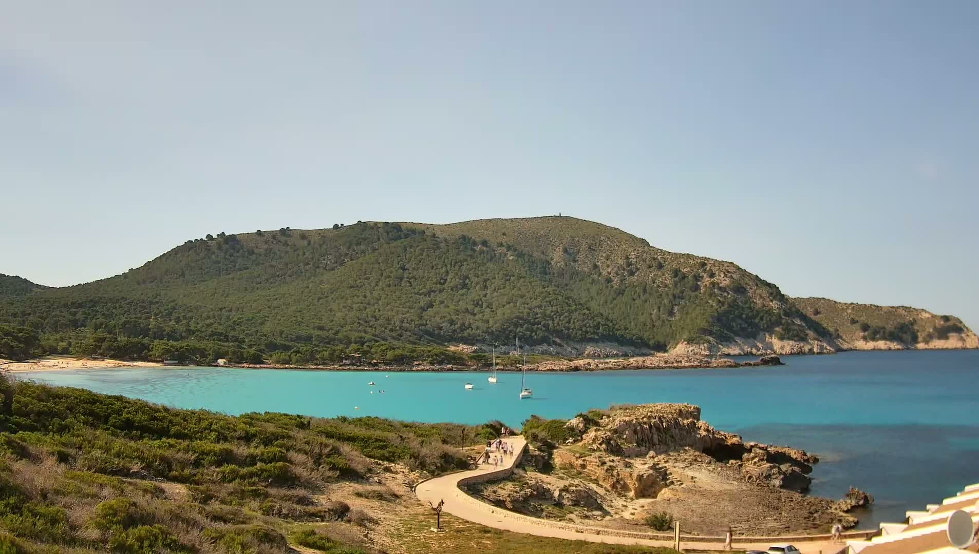 A curving path leads to a turquoise bay with several sailboats, a sandy beach, and a green, hilly landscape under a clear sky.