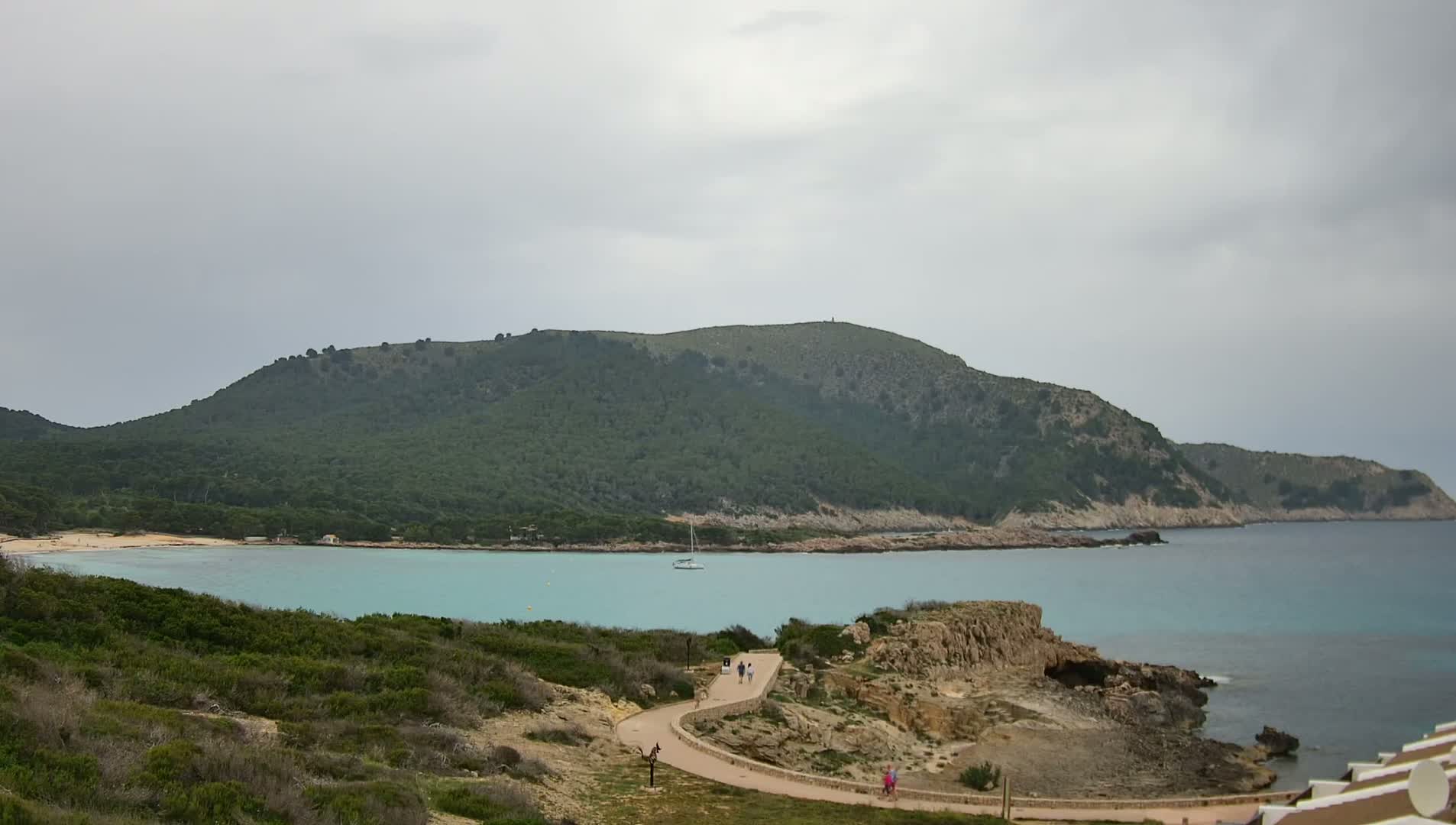 A winding path leads to a tranquil turquoise bay nestled between a verdant, hilly coastline under an overcast sky.