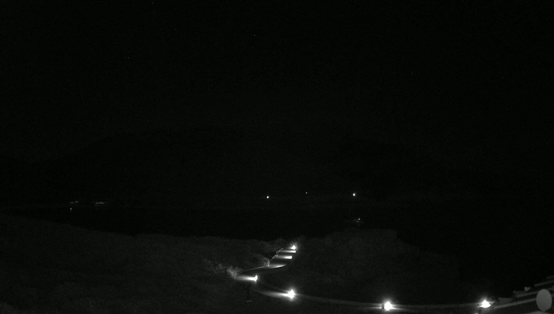 A dark, nighttime scene shows a winding road illuminated by lights, leading down toward a dark body of water under a clear, starlit sky.