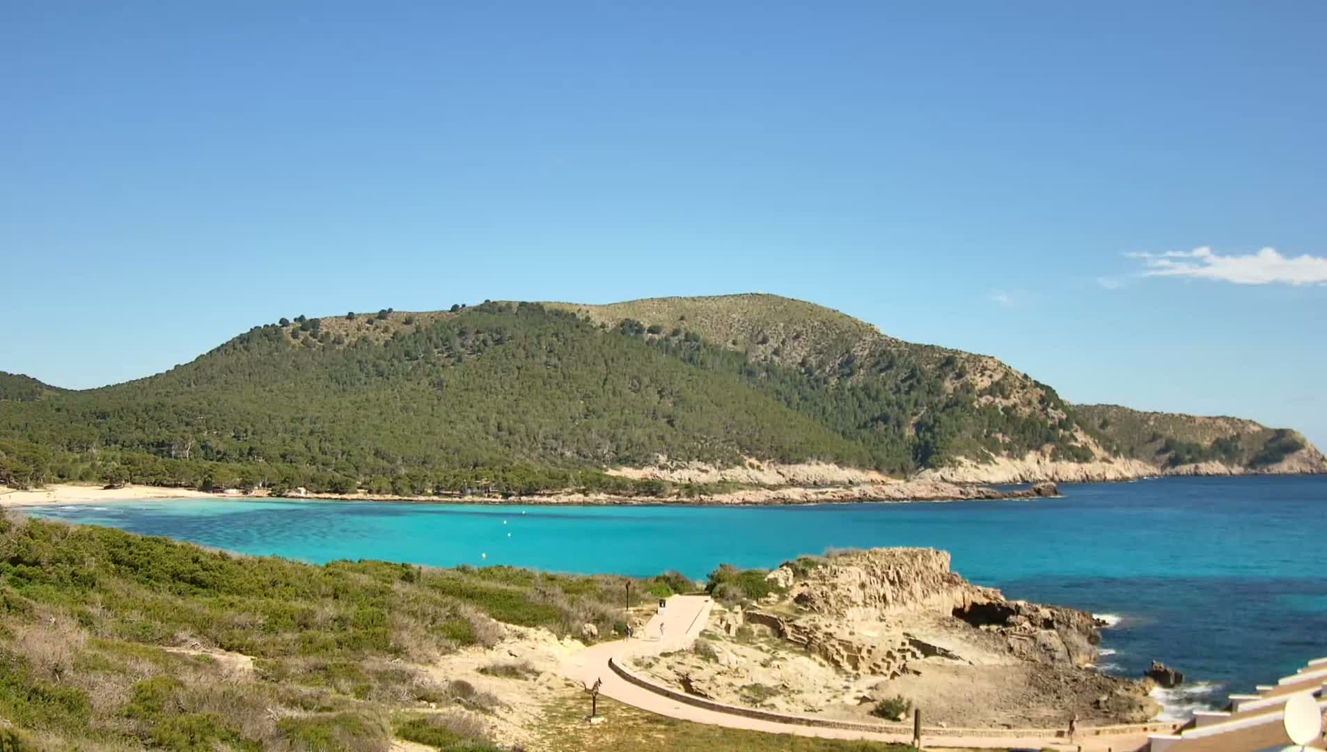 A sunny day reveals a secluded cove with turquoise water, a sandy beach, and a verdant, tree-covered hill in the background.