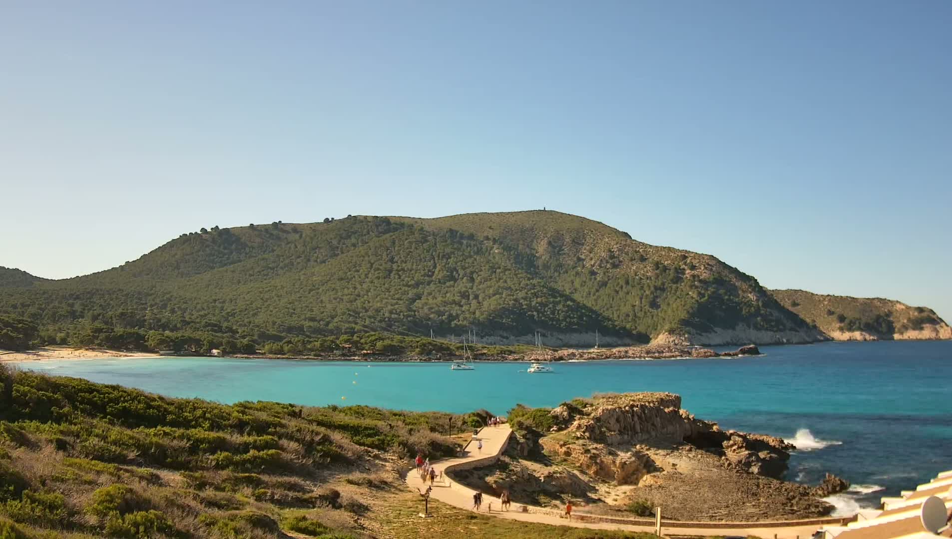 A tranquil turquoise bay with a sandy beach is nestled beneath a verdant, tree-covered hill on a sunny day.