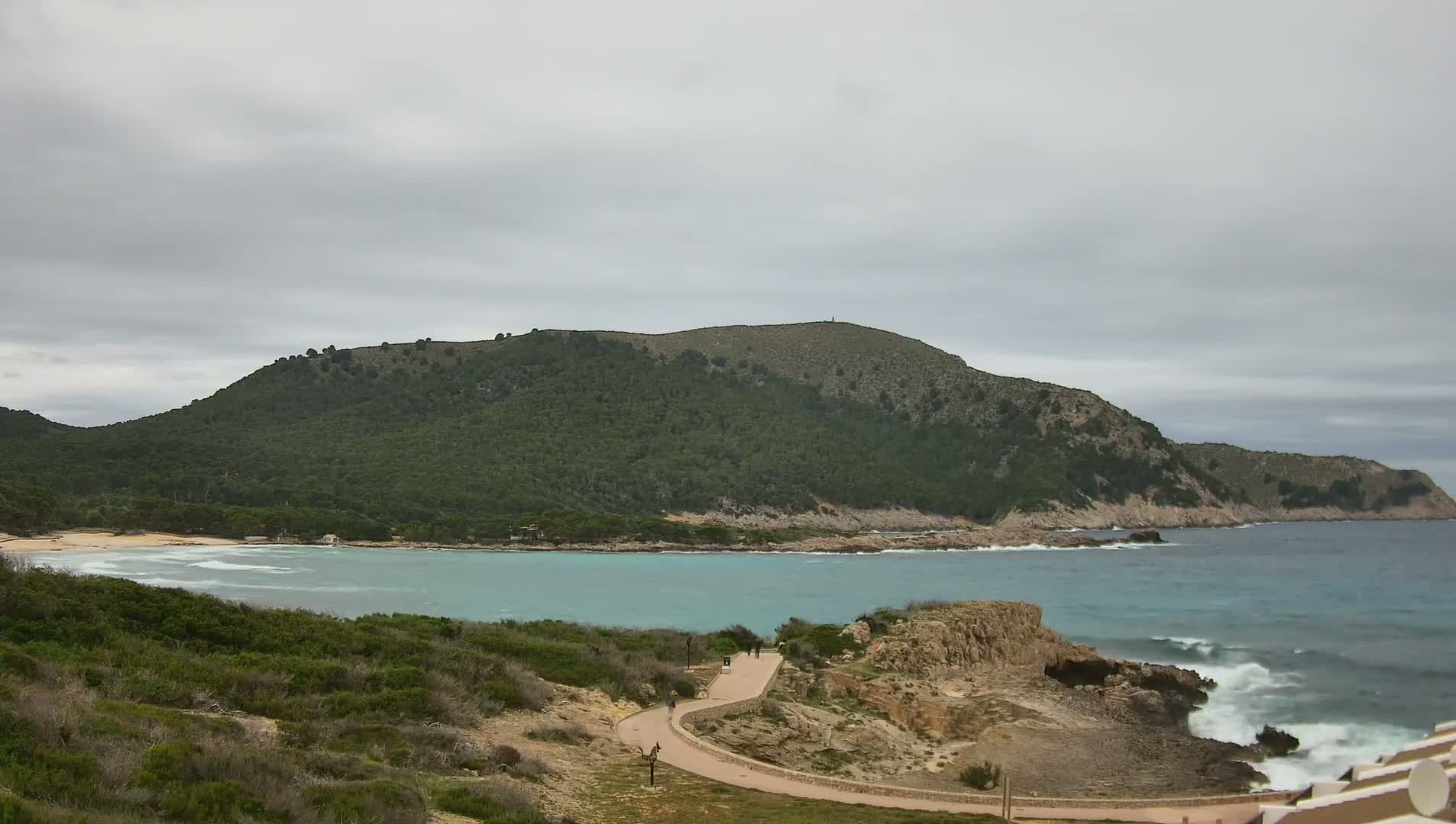 A winding path leads to a secluded beach nestled in a cove, bordered by a verdant, mountainous landscape under an overcast sky.