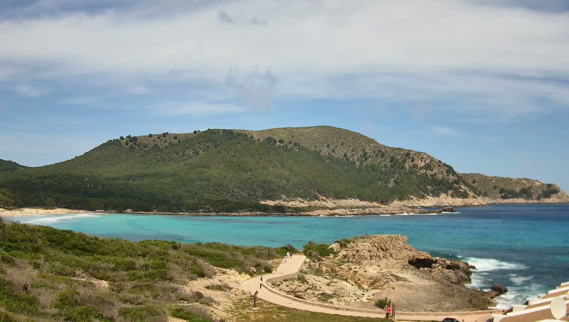 A paved path winds along a rocky coastline overlooking a turquoise bay with a sandy beach and a green hill in the background under a partly cloudy sky.