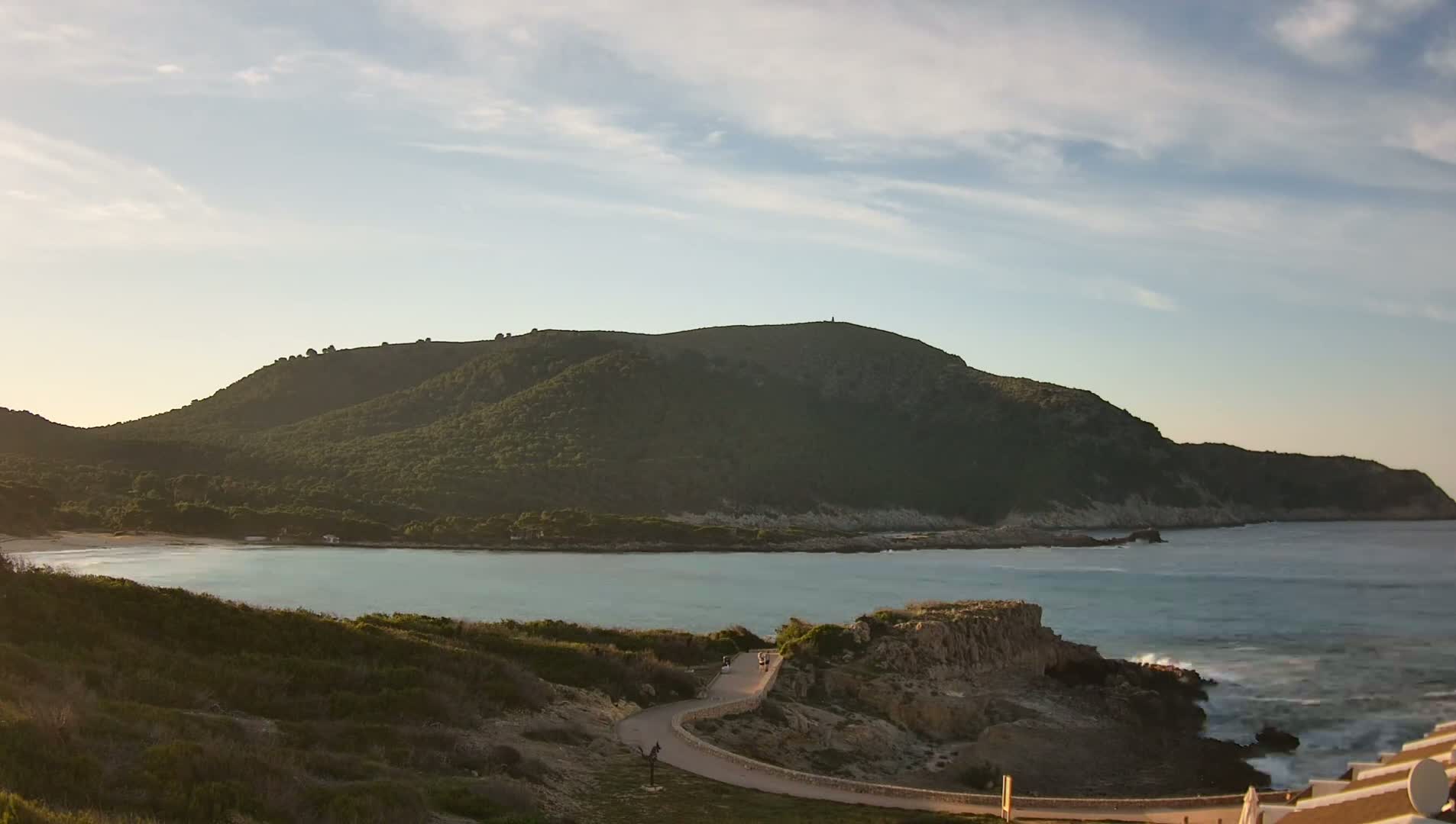 A tranquil coastal scene under a partly cloudy sky features a calm bay nestled between a verdant hillside and a rocky shoreline with gentle waves lapping the shore.
