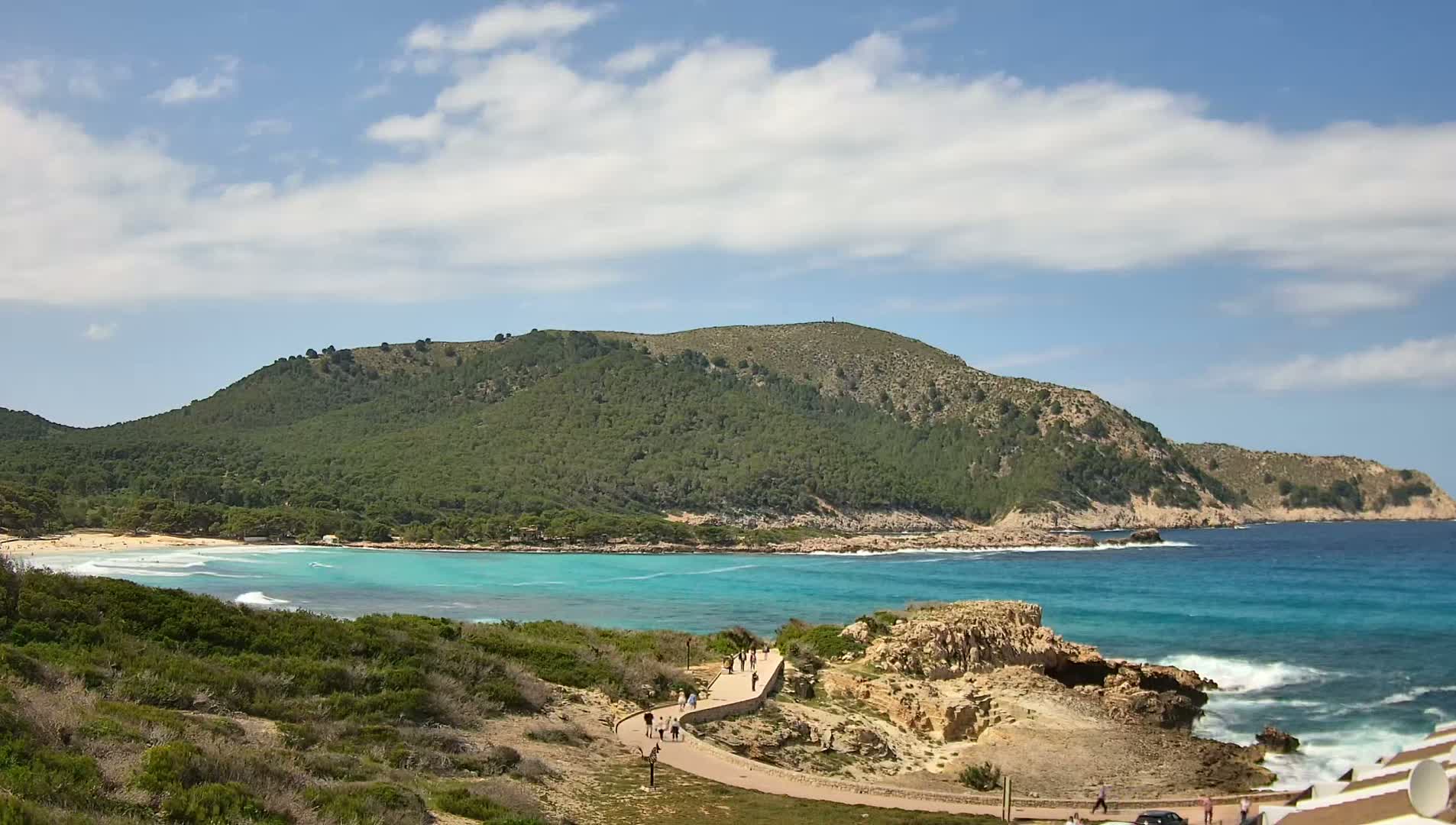 A paved path winds along a rocky coastline overlooking a turquoise bay with a sandy beach and a green, hilly landscape under a partly cloudy sky.