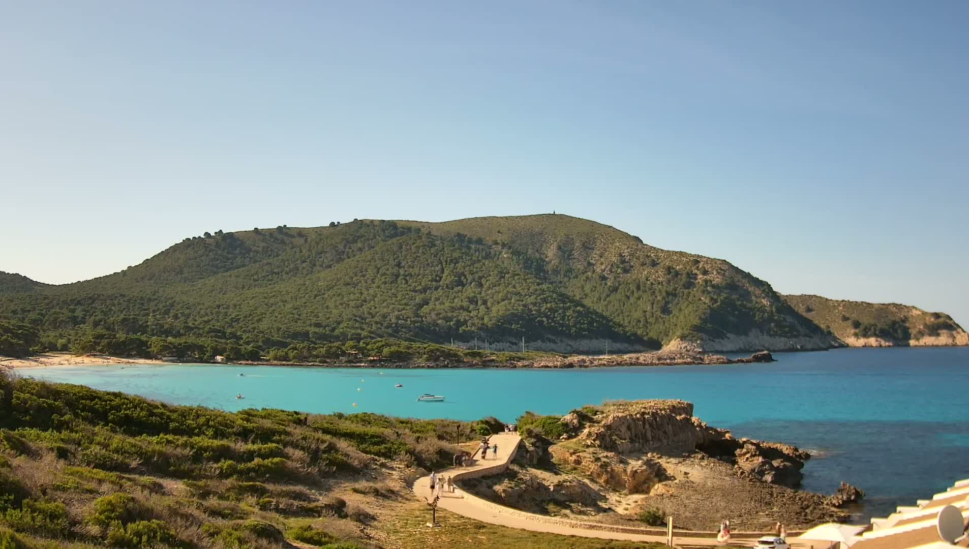 A turquoise bay with a sandy beach, nestled below a lush green hill, on a sunny day.