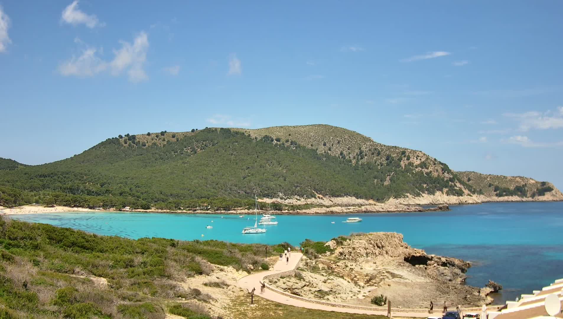A scenic coastal view on a sunny day shows a turquoise bay with several sailboats, a sandy beach, a green hill, and a paved path winding along the shoreline.