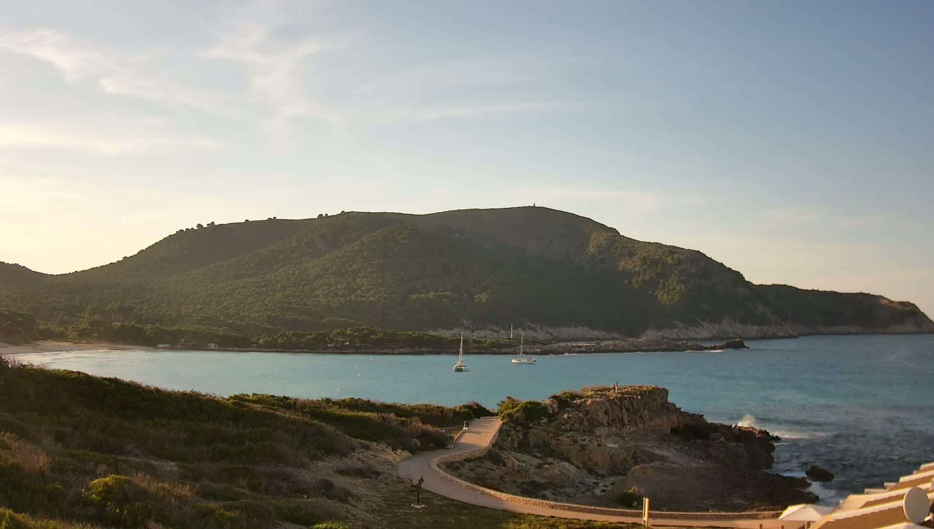 A tranquil coastal scene under a partly cloudy sky shows a verdant hill overlooking a calm bay with two sailboats, a sandy beach, and a winding path leading to the water's edge.