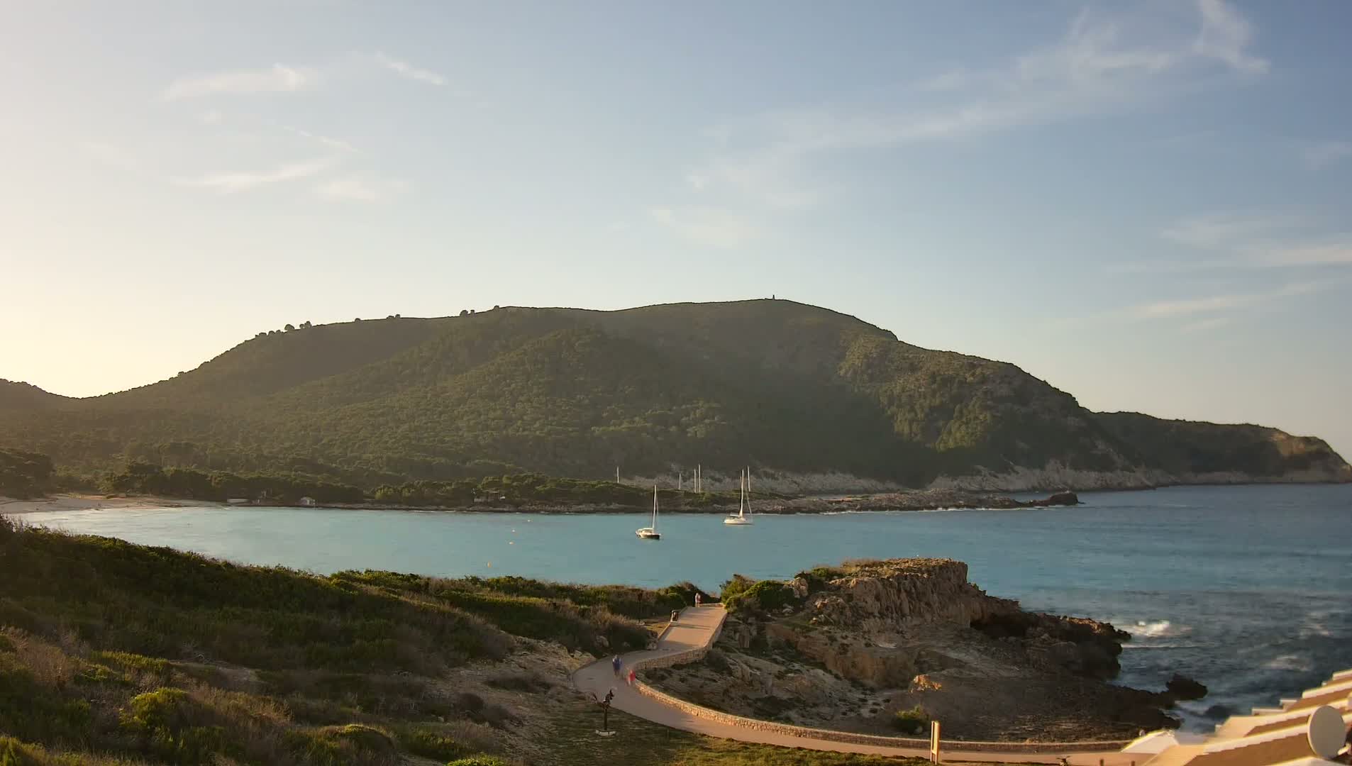 A tranquil coastal scene under a mostly clear sky shows a calm bay nestled between a verdant hill and rocky shoreline, with a few sailboats at anchor and people walking a path along the coast.