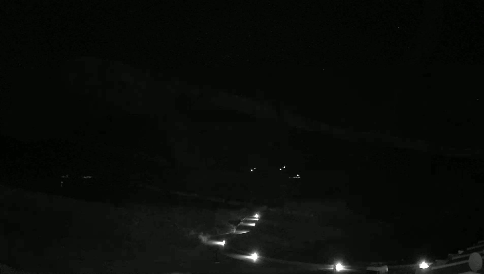 A nighttime, low-light view shows a winding road with several distant lights, possibly from vehicles, against a dark, mostly clear sky.